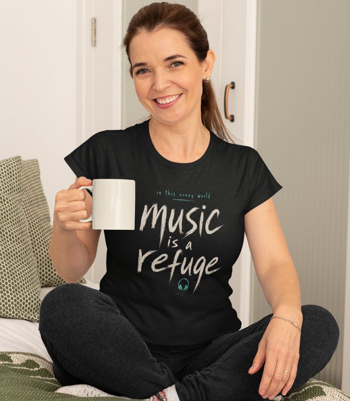A woman sitting cross-legged on a bed holding a white mug, wearing a black graphic t-shirt with the text 'music is a refuge' in white and gray lettering.
